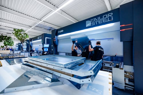 "Medewerkers van 247TailorSteel bewerken metaal, met stapels onderdelen op de voorgrond."